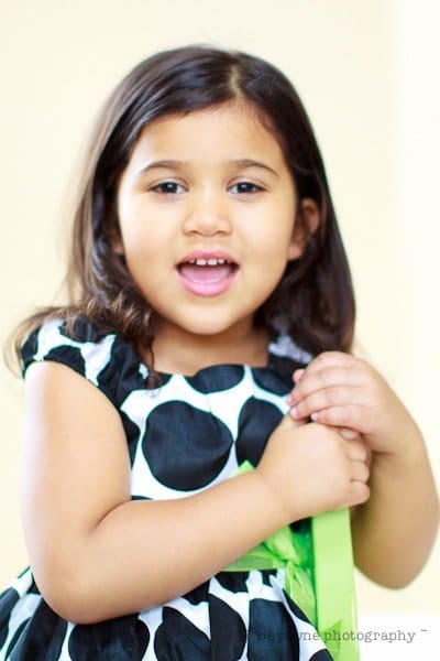 a star is born... A young girl with long, dark hair is smiling. She is wearing a black and white polka dot dress and holding onto something green. The background is softly blurred, creating a warm and cheerful atmosphere.