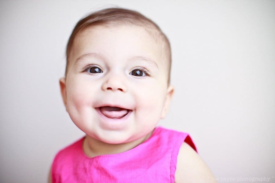 A smiling baby wearing a bright pink top against a plain background. The baby has short, dark hair and visible dimples on both cheeks.
