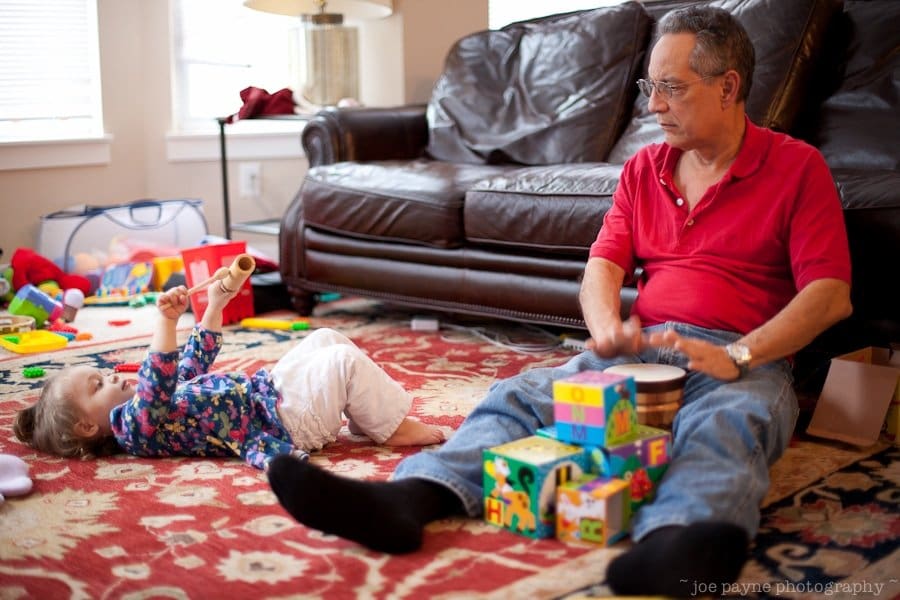 An older man in a red shirt sits on the floor playing a drum while a young child lies nearby, playing with a toy. Colorful building blocks surround them on a patterned rug in a cozy living room.