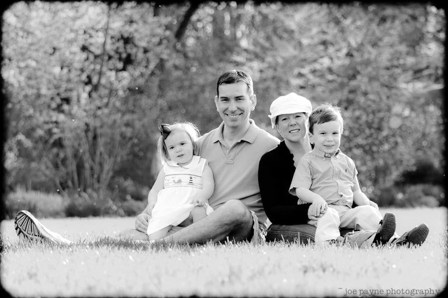 one sunday in spring... A black and white photo of a family sitting on grass in a park. Two adults and two young children are smiling at the camera. The adults are seated with the children on their laps. Trees and greenery are visible in the background.
