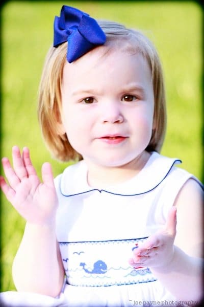 one sunday in spring... A young child with short hair wears a white dress with a blue design and a matching blue bow in their hair. The child smiles and holds their hands up slightly. They are outdoors with a blurred green background.