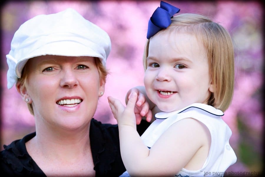one sunday in spring... A woman wearing a white cap holds a smiling toddler with a blue bow in her hair. Both are in front of a blurred background with pink hues.