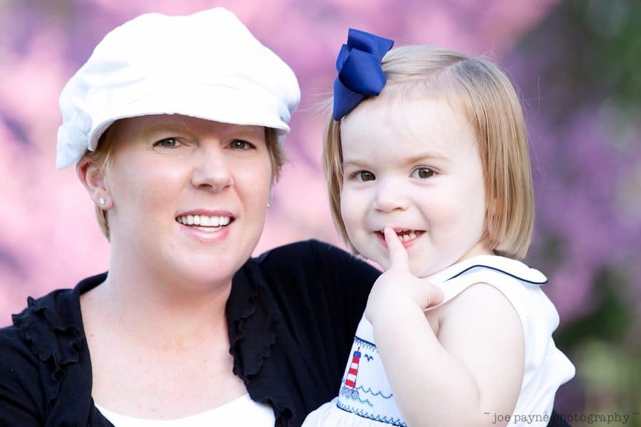 one sunday in spring... A woman wearing a white hat is smiling while holding a young child with a blue bow in her hair. The child is playfully touching her lips. The background is blurred with pink and purple hues.