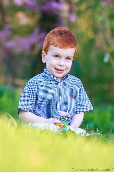 one sunday in spring... A young child with red hair, wearing a blue checkered shirt, sits on the grass holding a juice box. The background features blurred greenery and pink blossoms, creating a vibrant, springtime atmosphere.