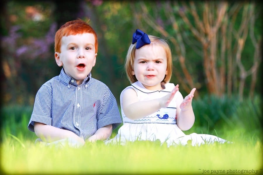 one sunday in spring... Two young children sit on the grass. The boy on the left has red hair and wears a checkered shirt. The girl on the right has light hair, a blue bow, and a white dress with blue trim, clapping her hands. They are outdoors with greenery in the background.