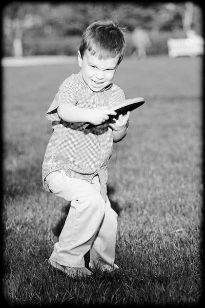 one sunday in spring... A young boy with a playful expression holds a frisbee, preparing to throw it. He is standing on grass, dressed in a checkered shirt and light pants. The black-and-white image captures the energetic moment in a park setting.