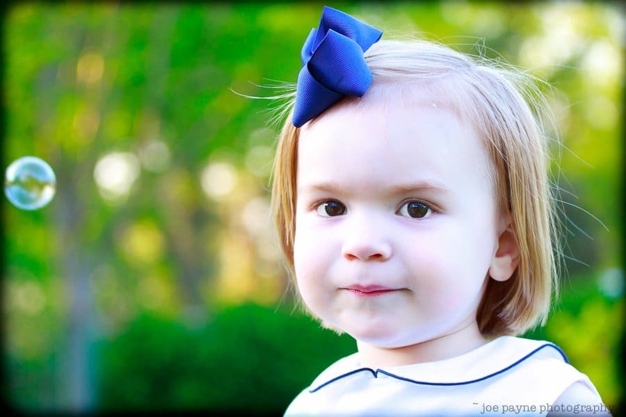 one sunday in spring... A young child with light brown hair and a blue bow on their head stands outdoors against a blurred green background, looking at a bubble floating nearby.