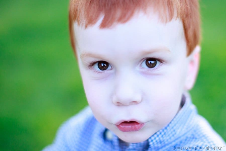 one sunday in spring... A close-up of a young child with red hair and brown eyes, wearing a blue checkered shirt. The child has a curious expression, and the background is a blurred green, possibly grass.
