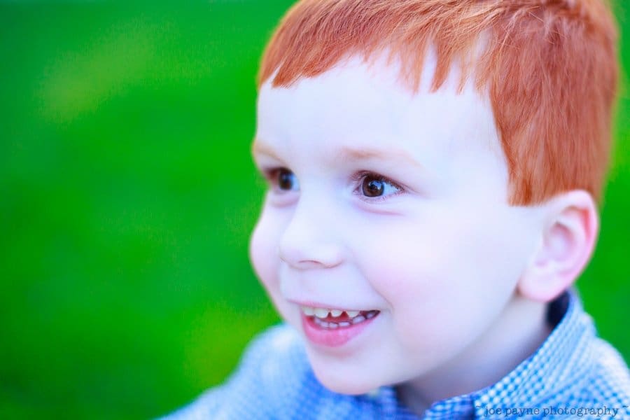 one sunday in spring... A young child with red hair is smiling and looking to the side. The background is a vibrant green, suggesting a grassy outdoor setting. The childs expression is joyful and curious, wearing a blue checkered shirt.