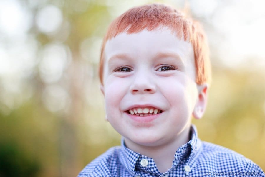 one sunday in spring... A young child with red hair smiles brightly at the camera, wearing a blue checkered shirt. The background is blurred, with hints of greenery and warm light.