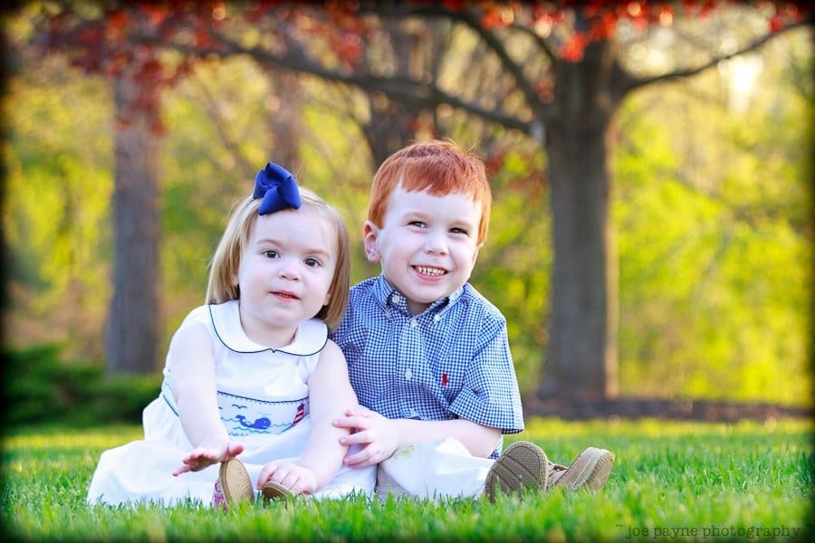 one sunday in spring... Two young children are sitting on the grass in a park. The girl is wearing a white dress with a blue bow, and the boy is in a checkered shirt. They are smiling, with trees and soft focus greenery in the background.