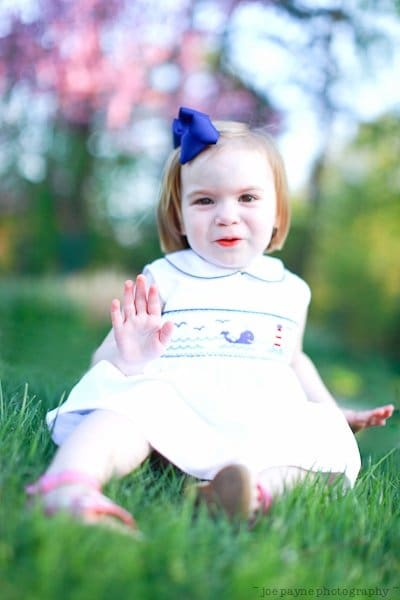 one sunday in spring... A young child with a blue bow in their hair sits on the grass, wearing a white dress with colorful stitching. The child is smiling and waving, with blurry trees and a pink flower in the background.