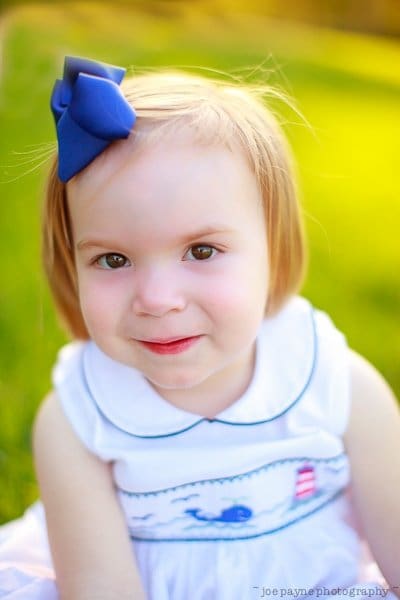 one sunday in spring... A young child with short hair and a blue bow smiles warmly at the camera, sitting on grass in a bright outdoor setting. She is wearing a white dress with blue trim.