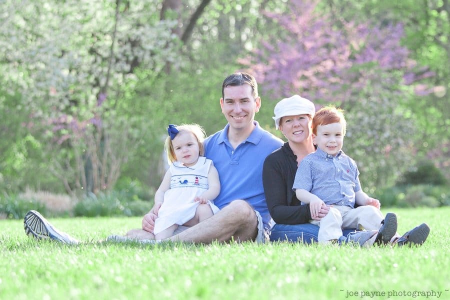 one sunday in spring... A family of four sits on lush green grass in a park. The father and mother are in the middle, with a young daughter in a white dress sitting beside the father and a young son in a blue shirt next to the mother. Blossoming trees are in the background.