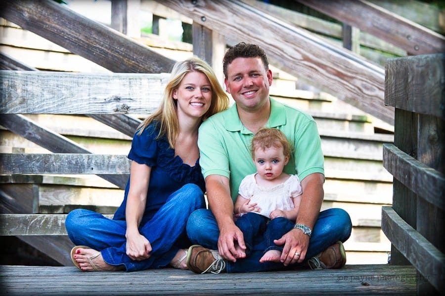 A smiling couple with a young child sitting together on a wooden bridge. The woman wears a navy blouse and jeans, the man wears a green polo shirt and jeans, and the child wears a white shirt and pink pants. The background is wooden railings and beams.