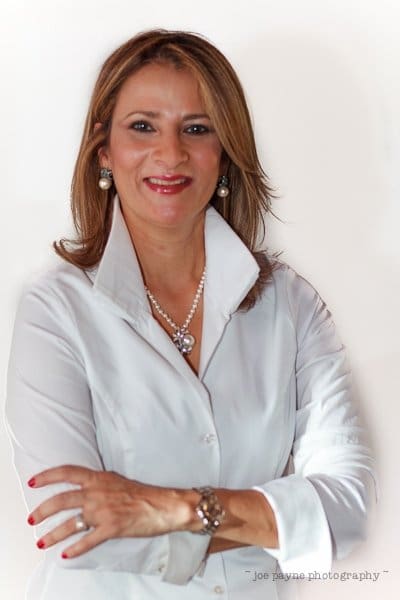 A confident woman with shoulder-length hair wearing a white blouse, pearl earrings, and necklace. She has her arms crossed and is smiling. The background is plain white.