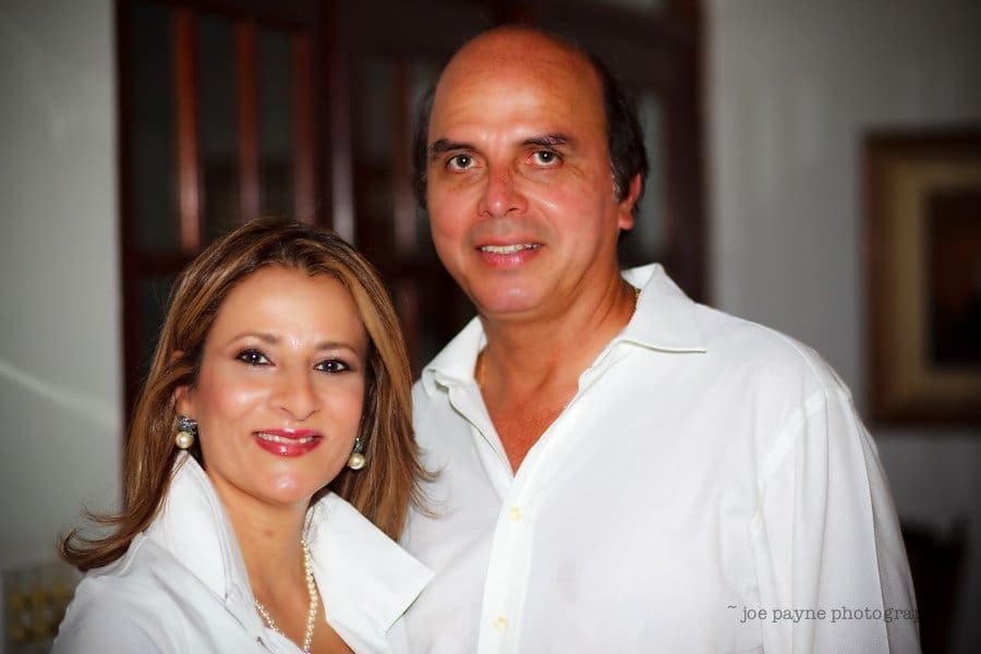 A smiling man and woman pose together indoors, both wearing white shirts. The woman has long hair and is wearing pearl earrings and a necklace. The man has short hair. The background is softly blurred.