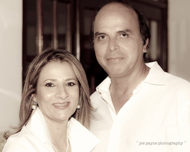 A woman and a man are standing together, both smiling. They are dressed in white shirts, and the background is indoors with dark wooden panels. The photo is in sepia tone.