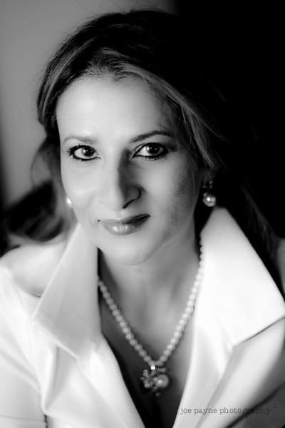 Black and white portrait of a woman with long hair, wearing a white blouse and pearl necklace, smiling at the camera.
