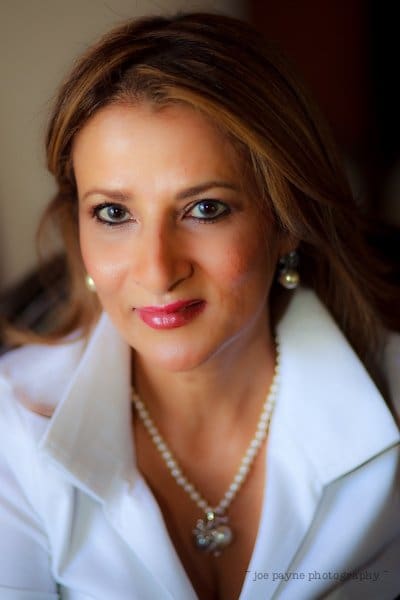 A woman with long hair wearing a white blouse and pearl necklace smiles softly at the camera. The background is softly blurred.