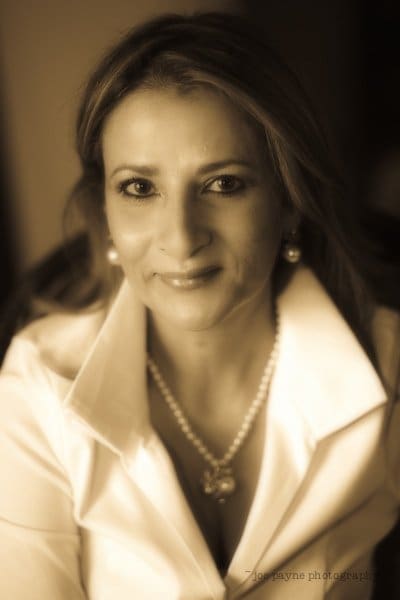 A woman with long hair, wearing a white blouse and pearl necklace, smiles gently at the camera. The image has a sepia tone, giving it a warm, vintage feel.