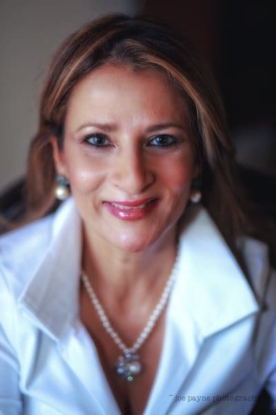 A woman with long hair smiles at the camera. She is wearing a white shirt and pearl earrings, with a necklace that has a pendant. The background is softly blurred.