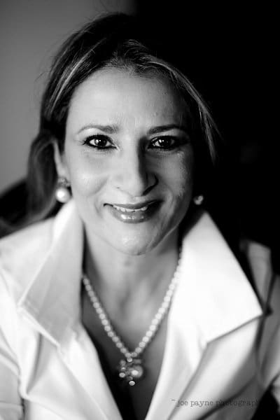 Black and white portrait of a person with long hair, smiling, wearing a white collared shirt and pearl necklace. The background is softly blurred to emphasize the subjects face.