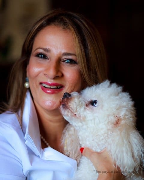 Woman smiling and holding a white fluffy dog close to her face. The dog appears to be nuzzling or sniffing her cheek. She is wearing a white blouse and has long brown hair. The background is dark and slightly blurred.