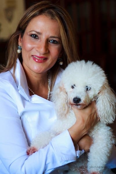 A woman with long hair smiles at the camera while holding a small white poodle. She is wearing a white shirt, and the background is softly blurred.