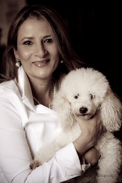 A woman with long hair, wearing a white blouse, smiles while holding a fluffy white poodle. The image is in sepia tones, giving it a warm, vintage feel.