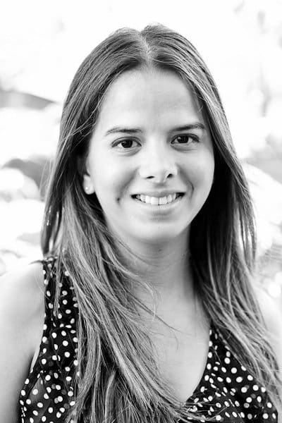 Black and white portrait of a young woman with long hair, smiling, wearing a polka dot top. The background is softly blurred.