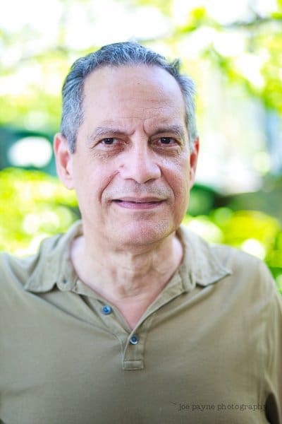 A man with short gray hair is wearing a beige shirt and standing outdoors. The background is blurred with greenery.