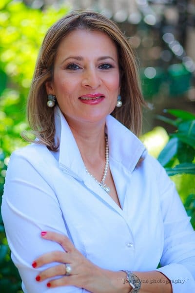 A woman with medium-length hair stands outdoors, smiling. She is wearing a white blouse and pearl earrings, with her arms crossed. The background is blurred greenery.