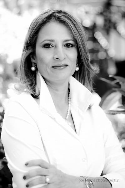 A black and white portrait of a person with shoulder-length hair, wearing a light-colored blouse and earrings. The background is softly blurred, emphasizing a warm, confident smile.