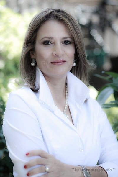 A woman with shoulder-length hair and wearing a white blouse is standing outdoors. She has crossed arms and is accessorized with pearl earrings and a necklace. The background is blurred with greenery.
