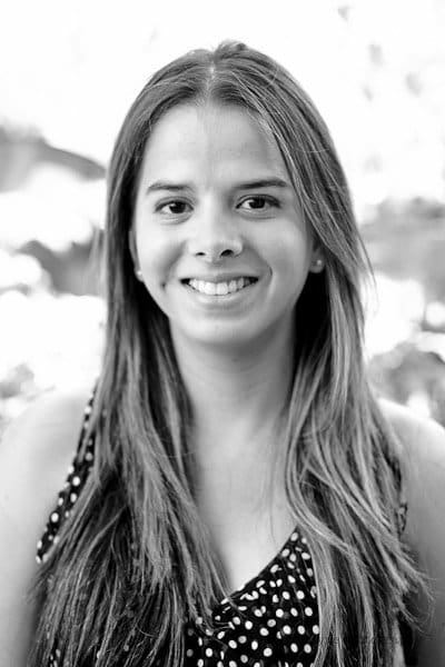 Black and white portrait of a smiling person with long hair, wearing a sleeveless polka dot top. The background is softly blurred.