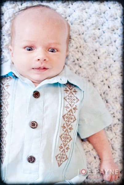 A baby lying on a textured white blanket, wearing a light blue outfit with decorative patterns and brown buttons.