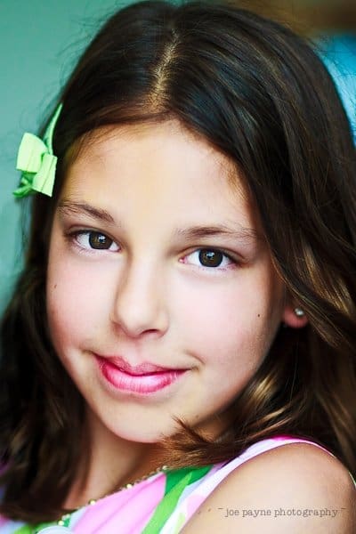 justin and meredith get hitched A young girl with brown hair smiles gently at the camera. She has a green bow in her hair and wears a colorful top. The background is softly blurred.