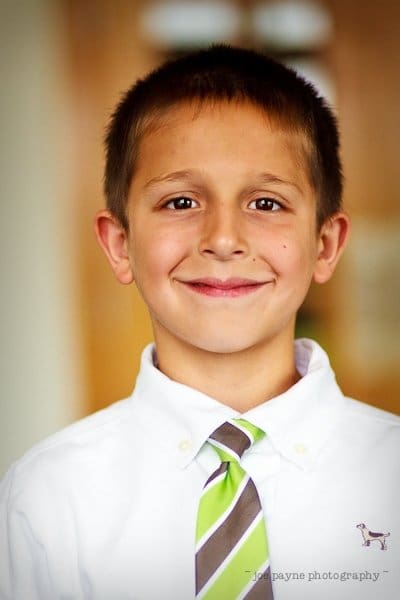 justin and meredith get hitched A young boy with short brown hair smiles, wearing a white shirt and a striped green and brown tie, standing in front of a blurred background.
