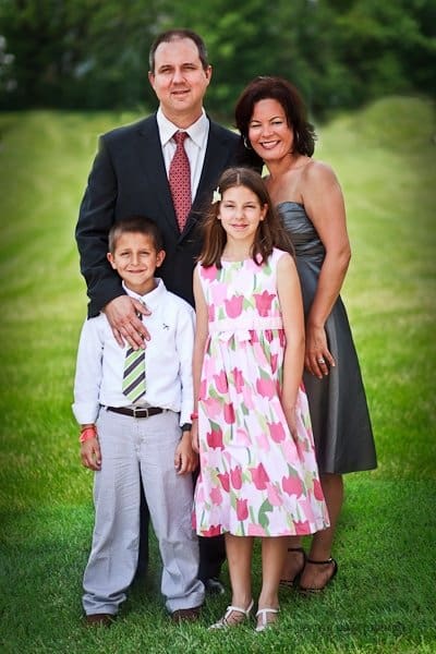 justin and meredith get hitched A family of four poses together on a grassy field. The father is in a suit, the mother in a gray dress, the son in a white shirt with a tie, and the daughter in a floral dress. They are smiling, with green trees in the background.