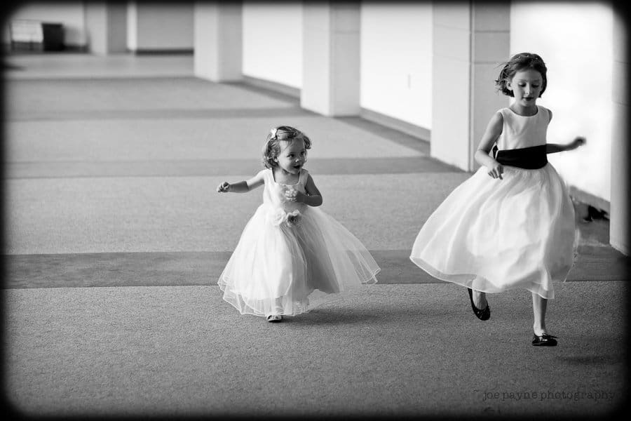 justin and meredith get hitched Two young girls in elegant dresses run happily down a carpeted hallway. One is slightly ahead, while the younger one follows, both wearing light-colored dresses with a gentle flow as they move. The scene is in black and white.