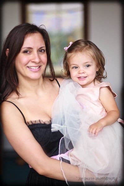 justin and meredith get hitched A woman in a black dress smiles while holding a young girl in a pink dress with a sheer overlay. Both have dark hair. The girl has a small pink bow in her hair. They are indoors with a framed picture in the background.