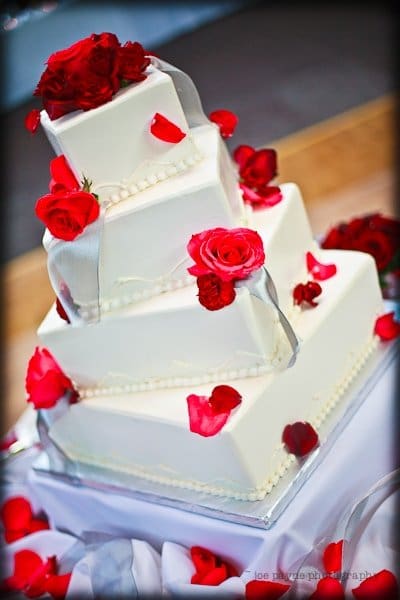 justin and meredith get hitched A tiered square wedding cake adorned with red roses and rose petals. Each layer is wrapped with a delicate white ribbon. The cake is set on a white tablecloth, creating an elegant and romantic display.