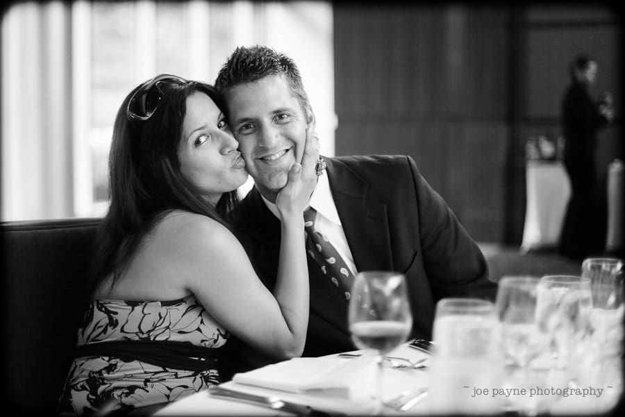 justin and meredith get hitched A woman playfully kisses a mans cheek as they sit at a table in a restaurant. He smiles at the camera, and she wears sunglasses on her head. The table is set for a meal, and the black-and-white image captures a joyful moment.