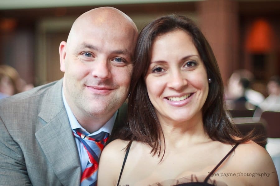 justin and meredith get hitched A smiling man in a gray suit with a loosened red tie sits close to a smiling woman in a black top. They are indoors with a blurred background of people and tables.