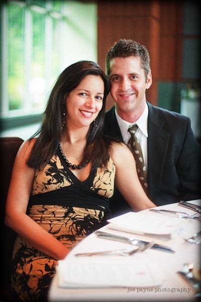 justin and meredith get hitched A smiling couple sits together at a restaurant table, with menus in front of them. The woman wears a black and tan dress, and the man is in a suit with a polka dot tie. They appear happy and relaxed.
