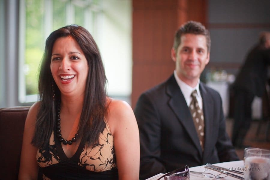 justin and meredith get hitched A woman with long dark hair is smiling, sitting beside a man in a suit with a patterned tie. They appear to be in a dining setting with blurred tables in the background and natural light coming through large windows.