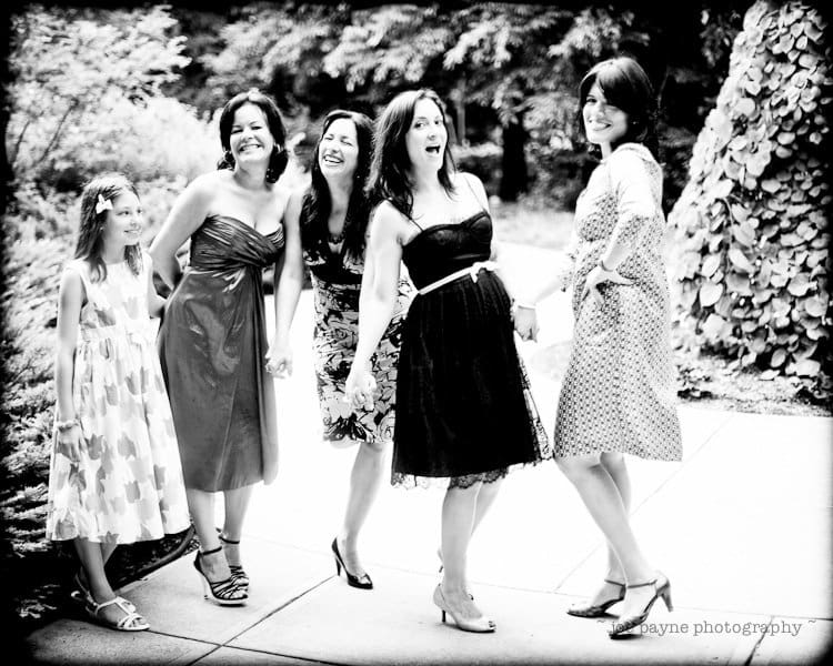 justin and meredith get hitched A black and white photo shows five women and a girl laughing and holding hands outdoors. They are dressed in elegant dresses, standing on a path with trees and greenery in the background.