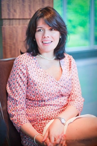 justin and meredith get hitched A woman with shoulder-length dark hair sits indoors, wearing a red and white patterned dress and a pearl necklace. She is smiling and looking slightly to the side. The background is softly blurred with a hint of greenery outside.
