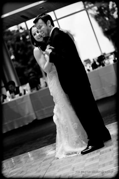 justin and meredith get hitched A black-and-white photo of a bride and groom smiling while dancing together at their wedding reception. They are surrounded by guests seated at tables in a softly lit venue. The back wall has large windows, revealing trees outside.